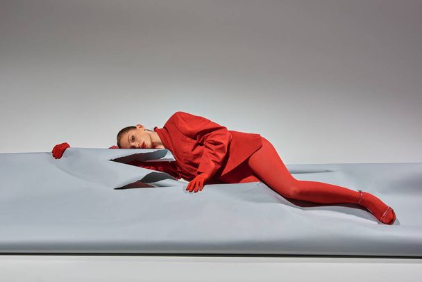молодая женщина в красной одежде в перчатках и колготках, лежащих рядом с разорванной бумагой на сером фоне - Фото, изображение