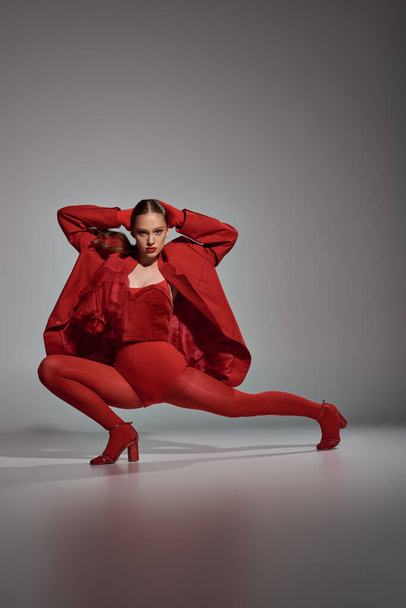 με αυτοπεποίθηση μοντέλο σε κομψό κόκκινο ντύσιμο με καλσόν και ψηλά τακούνια ποζάρουν σε γκρι φόντο - Φωτογραφία, εικόνα