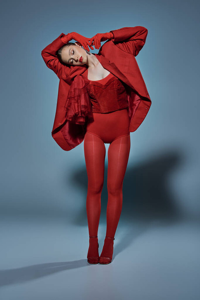 módní model se zavřenýma očima v červeném oblečení stojící v neobvyklé póze na šedém pozadí - Fotografie, Obrázek