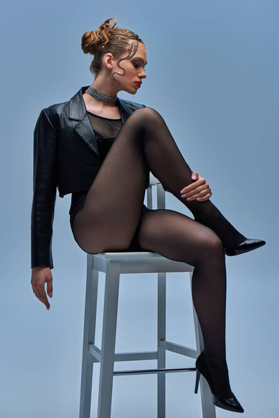 modelo de moda joven en chaqueta de cuero negro, medias y tacones altos sentado en la silla en el fondo gris - Foto, imagen