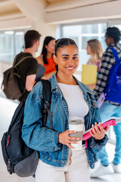 Üniversite kampüsünün koridorlarında elinde bir fincan kahve ve okul malzemesiyle arka planda çok ırklı sınıf arkadaşlarıyla kameraya bakan Latin öğrenci portresi.. - Fotoğraf, Görsel
