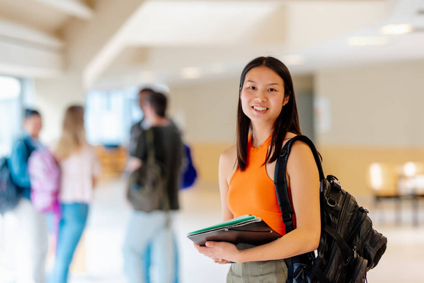 Portret Azjatki z plecakiem i zaopatrzeniem szkolnym na korytarzu kampusu uniwersyteckiego z kolegami z klasy w tle. - Zdjęcie, obraz