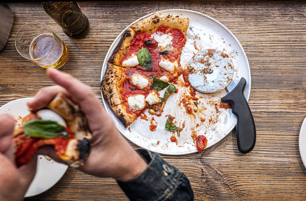 Мужская рука берет кусок свежеиспечённой неаполитанской пиццы Маргарита на деревенском деревянном столе в традиционной пиццерии. - Фото, изображение