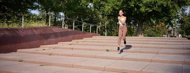 Zdravá fitness dívka běží venku na ulici, na sobě uniformu, běhání na čerstvém vzduchu a poslech hudby v bezdrátových sluchátkách. - Fotografie, Obrázek