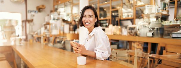 Portrét mladé brunetky, sedící s kávou a pomocí smartphonu v kavárně, chatování na mobilním telefonu. - Fotografie, Obrázek