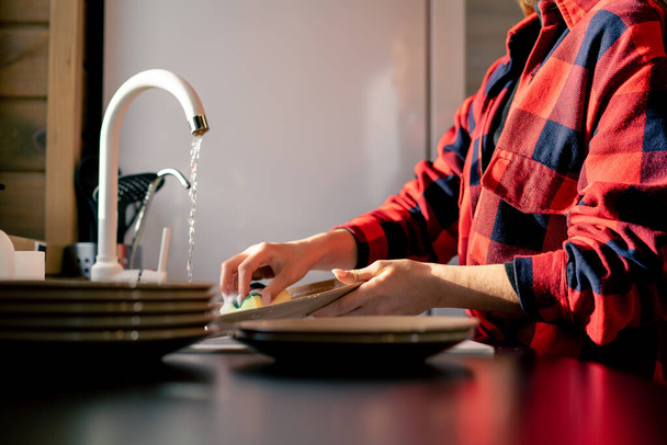 A girl in a checked shirt thoroughly washes dishes with foam in the kitchen of a wooden house - Foto, afbeelding