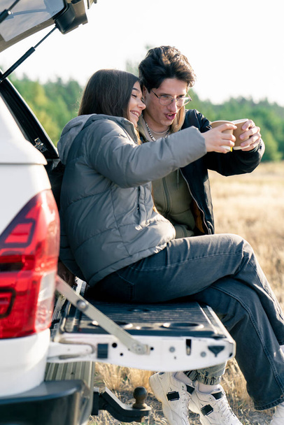 Egy pár megállt egy kocsiban egy mezőn, a csomagtartóban ültek és forró teát ittak egy termoszból az úton egy mezőn. - Fotó, kép