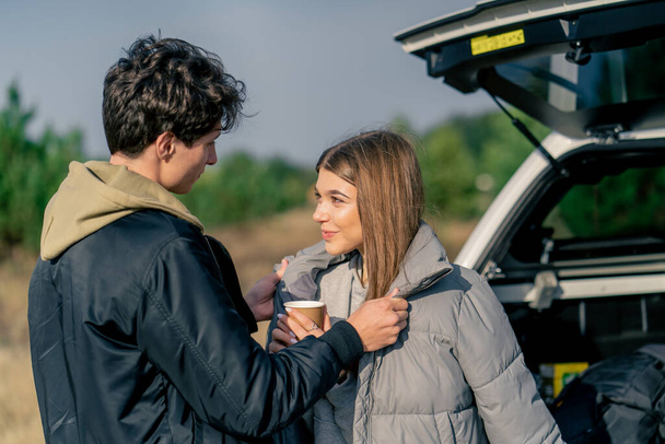 Um cara coloca um casaco em sua namorada com uma xícara de chá enquanto está perto do porta-malas aberto do carro na natureza - Foto, Imagem