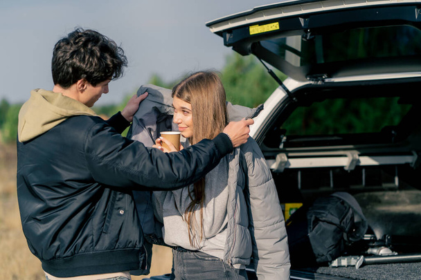 Парень надевает куртку на свою девушку с чашкой чая, стоя возле открытого багажника машины на природе - Фото, изображение