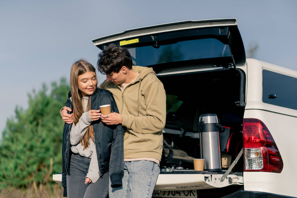 Ένας τύπος βάζει ένα σακάκι στην κοπέλα του με ένα φλιτζάνι τσάι ενώ στέκεται κοντά στο ανοιχτό πορτ-μπαγκάζ του αυτοκινήτου στη φύση - Φωτογραφία, εικόνα