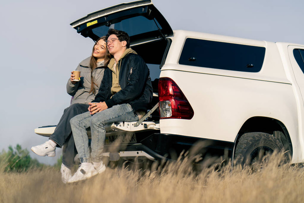 Um casal parou em um carro em um campo sentado no porta-malas e bebendo chá quente de uma garrafa térmica na estrada em um campo - Foto, Imagem