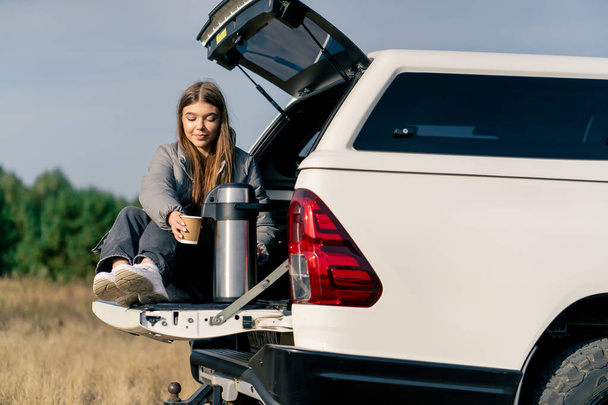 Piękna młoda dziewczyna w kurtce siedzi w bagażniku samochodu z termosem i pije gorącą herbatę na polu - Zdjęcie, obraz