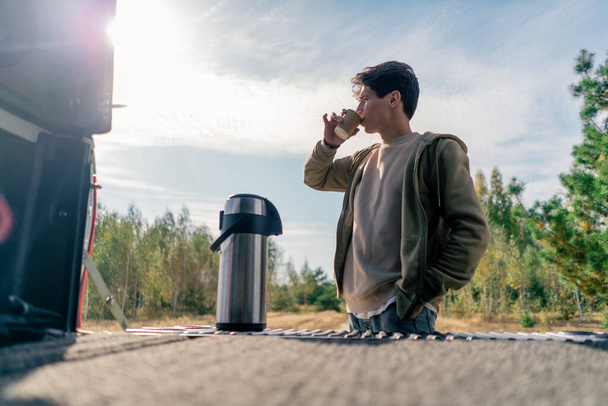 Um cara bebe um chá quente de uma garrafa térmica de metal grande, abrindo o porta-malas de seu carro enquanto viaja - Foto, Imagem