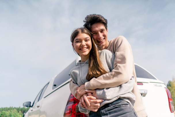 Portrait d'une jeune femme et d'un jeune homme riant et s'embrassant près de leur voiture en voyage en vacances - Photo, image