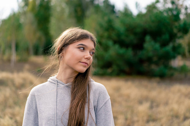 Portrait of a beautiful young girl with long hair standing in a field against background of pine trees - Photo, Image
