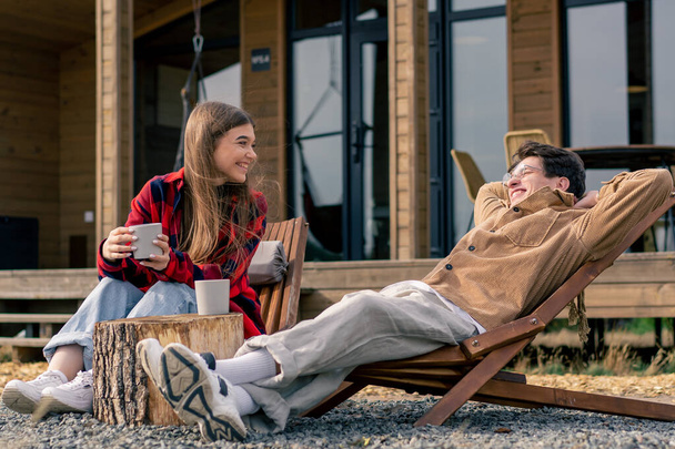 En la terraza cerca de una gran casa pareja enamorada se sienta con tazas de té en sillas de madera y se comunica entre sí - Foto, imagen