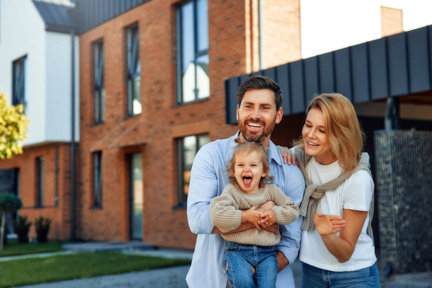 Moving of a young family to a new house. Couple with child standing in front of their newly bought house. Renting and buying a home. - Photo, Image