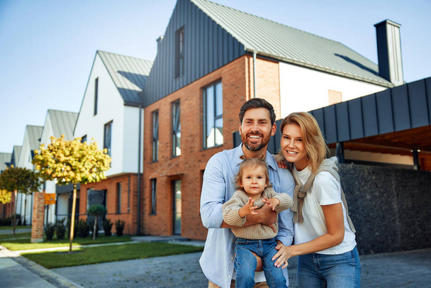 Moving of a young family to a new house. Couple with child standing in front of their newly bought house. Renting and buying a home. - Photo, Image