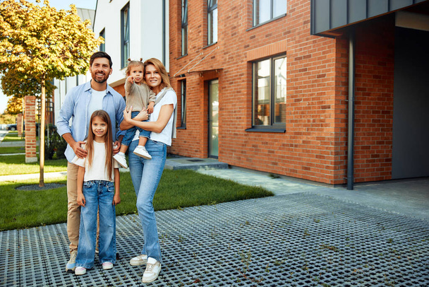Moving of a young family to a new house. Couple with two kids daughters standing in front of their newly bought house. Renting and buying a home. - Photo, Image