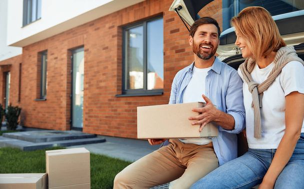 Young married couple sitting in the trunk of a car and unloading luggage in boxes to their new home. Renting and buying a home. Moving concept. - Photo, Image
