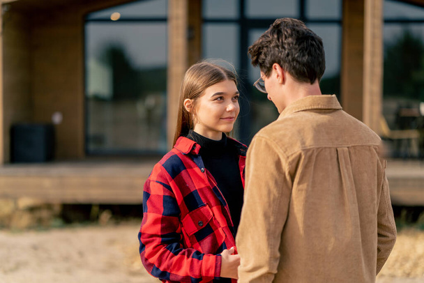 Una joven mujer mira a los ojos de su novio con amor y sonrisas mientras está de pie cerca de su casa de madera - Foto, imagen