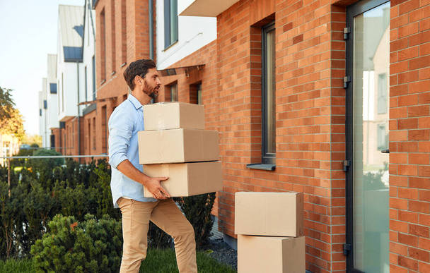 Un giovane uomo attraente con un mucchio di scatole in mano li porta nella sua nuova casa. Affittare e comprare una casa. Concetto in movimento. - Foto, immagini