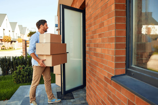 Un joven atractivo con un montón de cajas en sus manos las trae a su nuevo hogar. Alquilar y comprar una casa. Concepto móvil. - Foto, Imagen