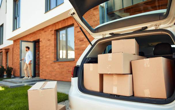 Ένα ανοιχτό πορτ-μπαγκάζ ενός αυτοκινήτου με ένα μάτσο κουτιά με υπάρχοντα, στο παρασκήνιο ένας άνθρωπος στέκεται στην πόρτα του νέου του σπιτιού. Νοικιάζοντας και αγοράζοντας ένα σπίτι. Μετακίνηση. - Φωτογραφία, εικόνα