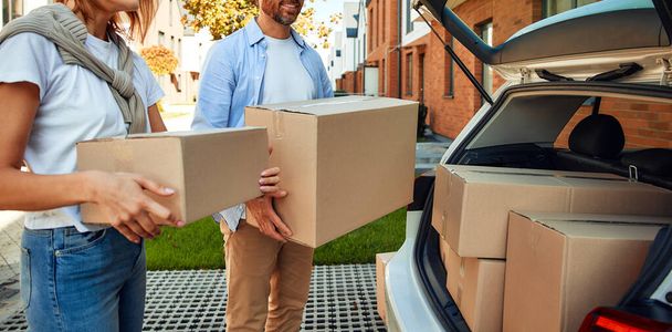 Ein junges Ehepaar lädt Kisten mit Dingen aus einem Auto und trägt sie in ein neues Zuhause. Miete und Kauf eines Eigenheims. Bewegendes Konzept. - Foto, Bild