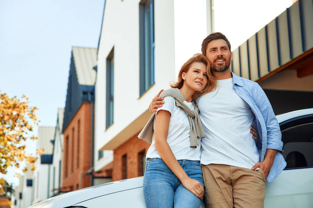 Een jong getrouwd stel dat naast een auto staat, geparkeerd bij een nieuw huis. Een huis huren en kopen. Bewegend concept. - Foto, afbeelding
