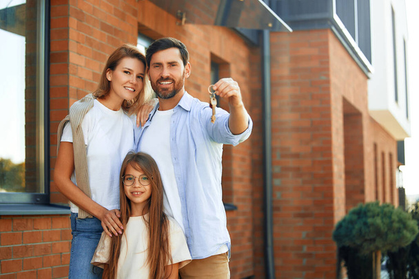 Eine junge Familie mit ihrer Tochter steht vor ihrem neuen Haus und zeigt die Schlüssel dazu. Miete und Kauf eines Eigenheims. Bewegendes Konzept. - Foto, Bild
