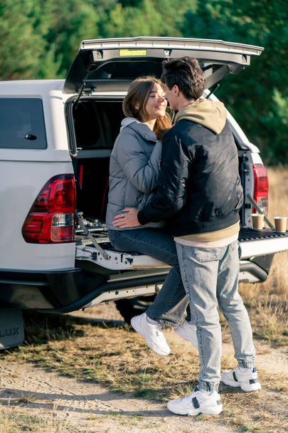A young couple in love a guy and a girl are resting together in the trunk of car hugging and kissing outdoors in a field - Photo, Image