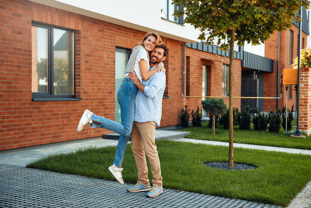 Una giovane coppia di sposi felici in piedi vicino a una nuova casa e abbracciati. Affitto e acquisto di immobili. Concetto in movimento. - Foto, immagini