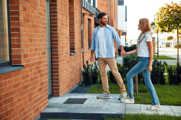 Una joven pareja casada feliz de pie cerca de una nueva casa y abrazándose. Alquilar y comprar bienes raíces. Concepto móvil. - Foto, imagen