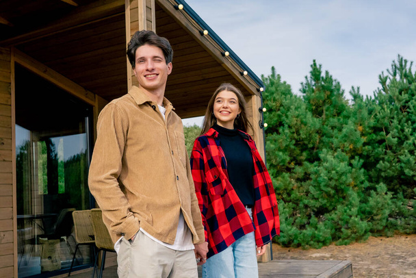 Giovane ragazzo e ragazza innamorati che si tengono per mano escono sulla terrazza di una casa in legno per passeggiare nella natura - Foto, immagini