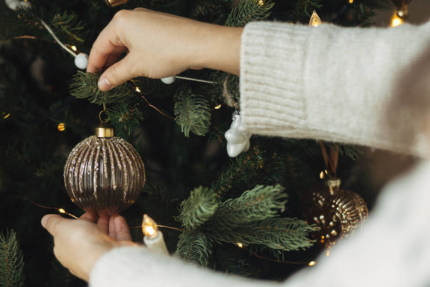 Hände in kuscheligen Pullovern hängen in Großaufnahme an Baumzweigen. Weihnachtsbaum mit stilvoller Christbaumkugel dekorieren. Atmosphärische Winterurlaubstradition, Familienzeit - Foto, Bild