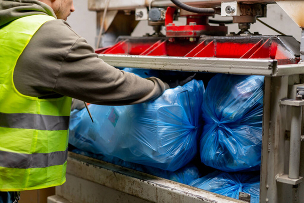 Κοντινό πλάνο αρσενικών χεριών σε γάντια που φορτώνουν μεγάλες σακούλες σκουπιδιών σε ειδική μηχανή συμπίεσης απορριμμάτων για διάθεση αποβλήτων - Φωτογραφία, εικόνα