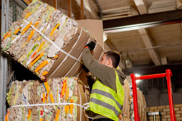 Egy férfi munkás védőmellényben és kesztyűben hatalmas papírdobozokat pakol egymásra egy gyárfolyosón. - Fotó, kép