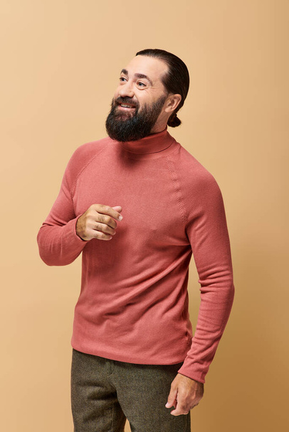 hombre feliz con barba en jersey de cuello alto rosa sonriendo alegremente sobre fondo beige - Foto, Imagen