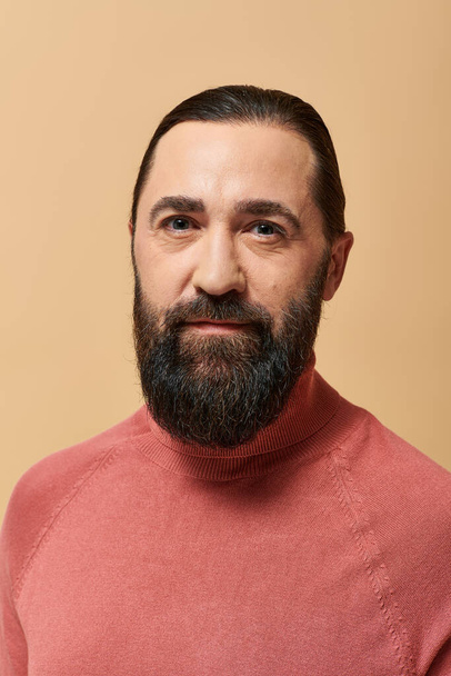 portrait, serious and handsome man with beard posing in pink turtleneck jumper on beige background - Photo, Image