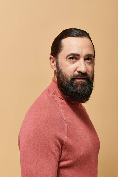 portrait, serious handsome man with beard posing in pink turtleneck jumper on beige background - Photo, Image