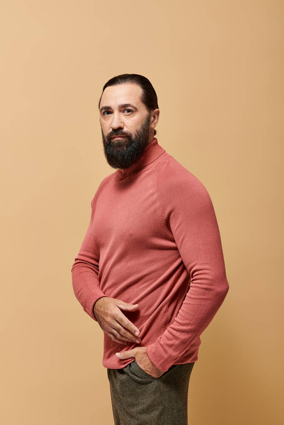 портрет, сосредоточенный и красивый мужчина с бородой позирует в розовой водолазке свитер на бежевом фоне - Фото, изображение