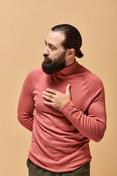 portrait of serious and handsome man with beard posing in pink turtleneck jumper on beige background - Photo, Image