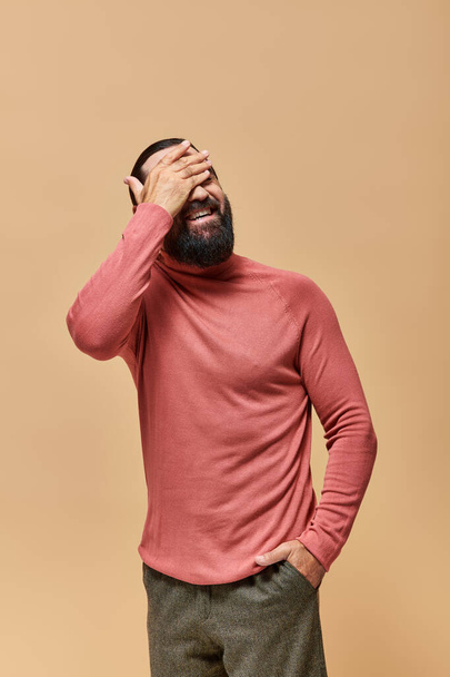 happy man with beard in pink turtleneck jumper smiling cheerfully on beige backdrop - Foto, afbeelding