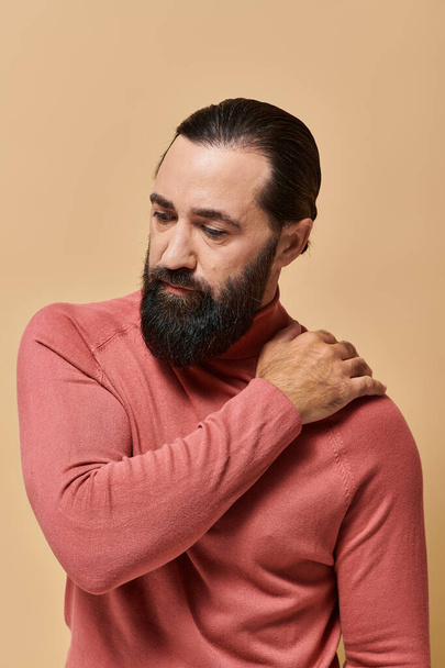 портрет, серьезный красивый мужчина с бородой позирует в розовой водолазке свитер на бежевом фоне - Фото, изображение
