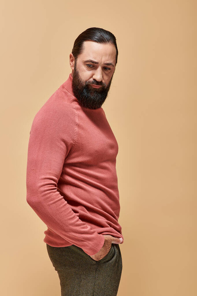 portrait, modèle sérieux et beau avec barbe posant en pull col roulé rose sur fond beige - Photo, image