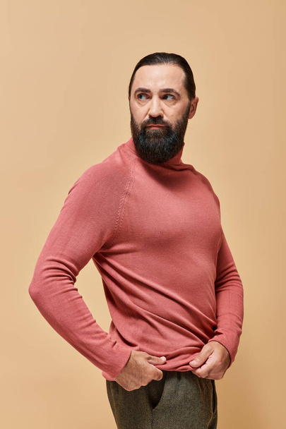 портрет, серйозний і красивий чоловік з бородою позує в рожевому стрибунці черепашки на бежевому фоні - Фото, зображення
