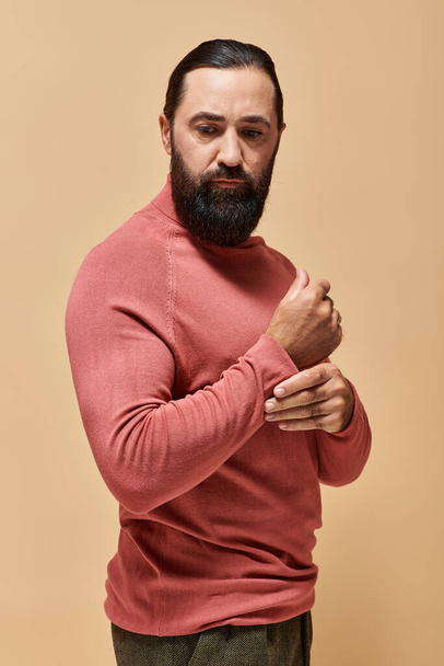 bel homme sérieux avec barbe posant en pull à col roulé rose sur fond beige, portrait - Photo, image