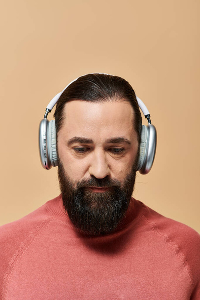 красивий бородатий чоловік в рожевому стрибку з черепашкою слухає музику в бездротових навушниках на бежевому - Фото, зображення