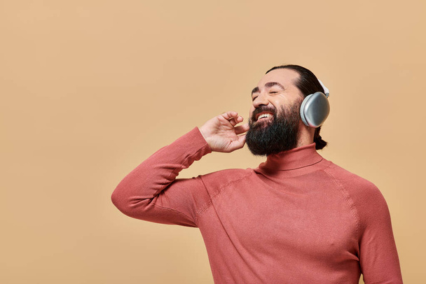 alegre hombre barbudo en jersey de cuello alto escuchando música en auriculares inalámbricos, sonriendo en beige - Foto, imagen